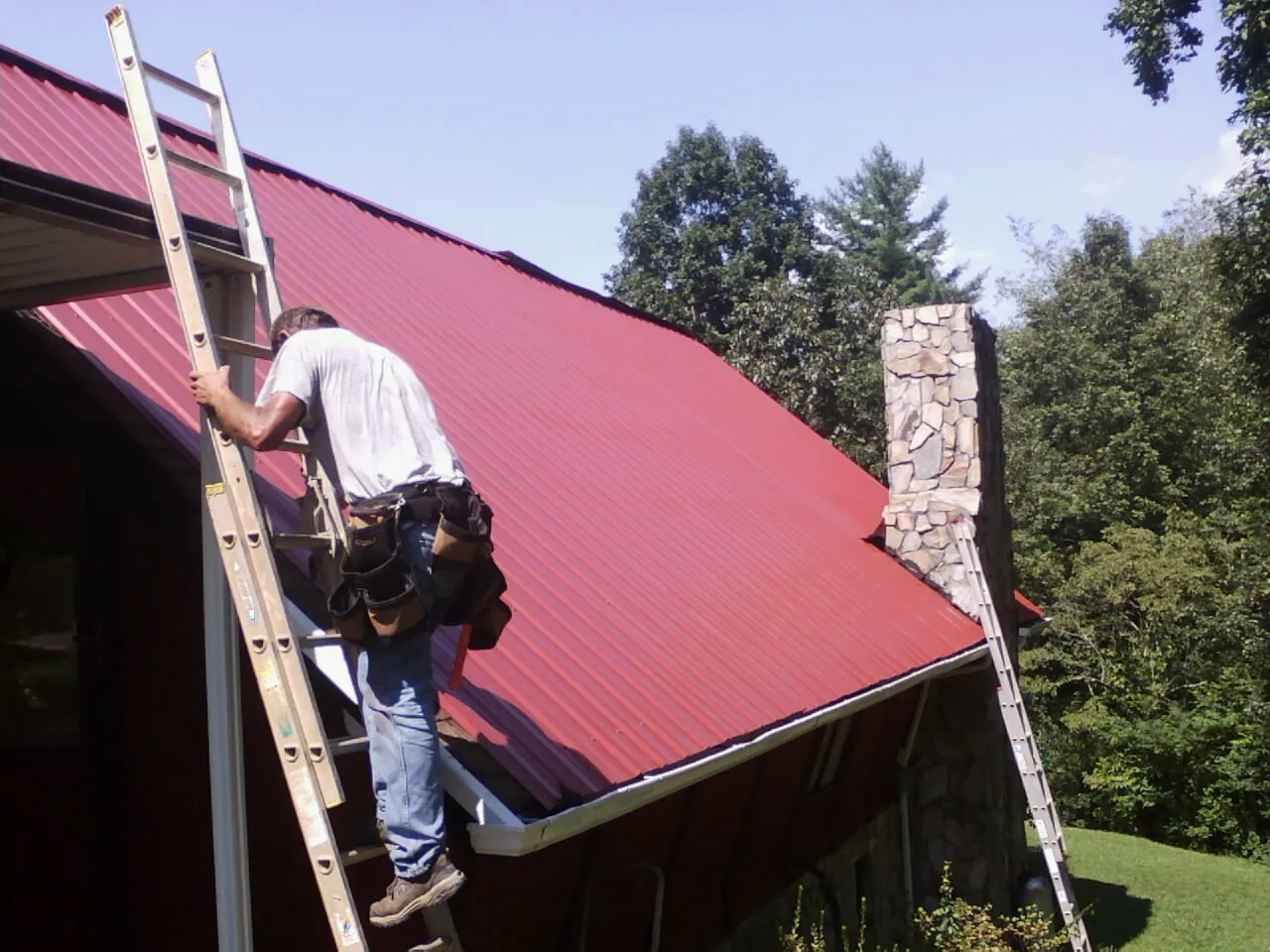 Roofer on a ladder beside a red metal roof and stone chimney — Soffit & Fascia Repair by Roofing Brothers in Haddon Heights
